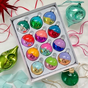 May include: Assortment of 12 colorful glass ornaments in various shades of blue, green, red, and purple. The ornaments are round and have a shiny finish. They are arranged in a white box with a white background.