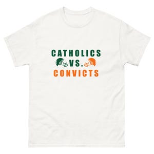 Puede incluir: Camiseta blanca con texto verde y naranja que dice "Catholics vs. Convicts" con dos cascos de fútbol a cada lado del "vs."