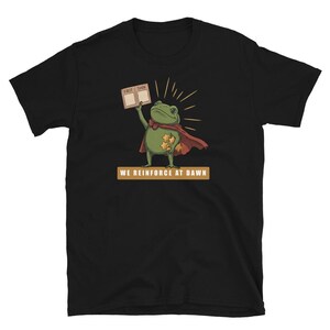 Könnte beinhalten: Ein schwarzes T-Shirt mit kurzen Ärmeln, das einen grünen Frosch mit rotem Umhang zeigt, der ein Schild mit der Aufschrift FIRST THEN und goldene Sterne hält. Unter dem Frosch steht auf einem orangefarbenen Banner WE REINFORCE AT DAWN.