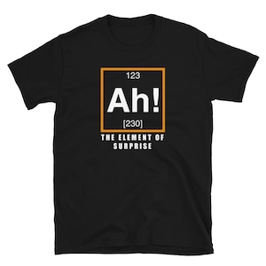 May include: Black t-shirt featuring a white and orange graphic. The design mimics a periodic table element, with "Ah!" in a square, and the words "THE ELEMENT OF SURPRISE" beneath it.