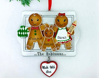 May include: A silver gingerbread family Christmas ornament with a red heart charm that says "Made With Love". The ornament features three gingerbread people with the names John, Ashley, and Sarah. The ornament also includes the year 2019 and the text "...The Robinsons..."