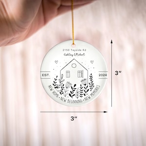 May include: A round white ceramic ornament measuring 3 inches by 3 inches, featuring a black line drawing of a house surrounded by flowers, hearts, and the text 2150 Tayside Rd, Ashley & Robert, EST. 2024, NEW HOME NEW BEGINNING NEW MEMORIES.