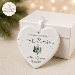 May include: A white heart-shaped Christmas ornament with the text "OUR FIRST CHRISTMAS AS mr & mrs" and the names "Vitor & Bianca" with the date "12.09.2026". The ornament has a white ribbon for hanging.