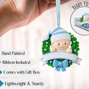 May include: Hand-painted Christmas ornament featuring a baby wearing a blue and white striped hat peeking out of a green holly wreath with a blue bow. The ornament comes with a ribbon and a gift box. "Ready to Gift" is written in a circle around the ornament.