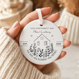 May include: A round white ceramic ornament featuring a black line drawing of a house surrounded by flowers. The text reads 3325 NORTHWEST AVE, Alex & Lindsay, EST. 2026, and NEW HOME NEW BEGINNING NEW MEMORIES. A white ribbon is attached to the top.