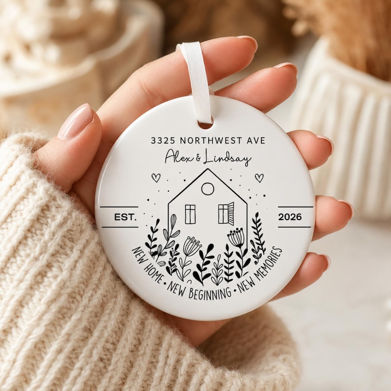 May include: A round white ceramic ornament featuring a black line drawing of a house surrounded by flowers. The text reads 3325 NORTHWEST AVE, Alex & Lindsay, EST. 2026, and NEW HOME NEW BEGINNING NEW MEMORIES. A white ribbon is attached to the top.