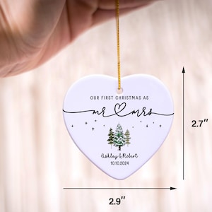 Puede incluir: Adorno navide&ntilde;o de cer&aacute;mica blanca en forma de coraz&oacute;n con el texto "Our First Christmas as Mr & Mrs Ashley & Robert 10.10.2024" y una ilustraci&oacute;n de un pino verde. El adorno mide 2,9 pulgadas de ancho y 2,7 pulgadas de alto.