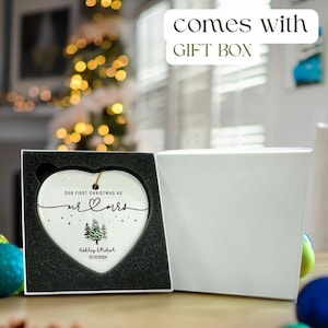 Puede incluir: Un adorno de cer&aacute;mica blanco en forma de coraz&oacute;n con el texto "Our first Christmas as Mr & Mrs" y un peque&ntilde;o dise&ntilde;o de coraz&oacute;n. El adorno tambi&eacute;n presenta una ilustraci&oacute;n de acuarela de dos pinos y el texto "Ashley & Robert 10.10.2024". El adorno se muestra en una caja de regalo blanca con interior negro.