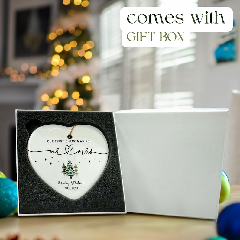 Puede incluir: Un adorno de cer&aacute;mica blanco en forma de coraz&oacute;n con el texto "Our first Christmas as Mr & Mrs" y un peque&ntilde;o dise&ntilde;o de coraz&oacute;n. El adorno tambi&eacute;n presenta una ilustraci&oacute;n de acuarela de dos pinos y el texto "Ashley & Robert 10.10.2024". El adorno se muestra en una caja de regalo blanca con interior negro.