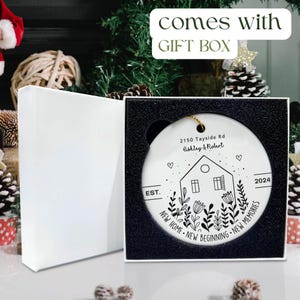 May include: A round white ceramic ornament featuring a black line drawing of a house, flowers, and the text 2150 Tayside Rd, Ashley & Robert, EST. 2024, and NEW HOME NEW BEGINNING NEW MEMORIES, presented in a white gift box with black foam.