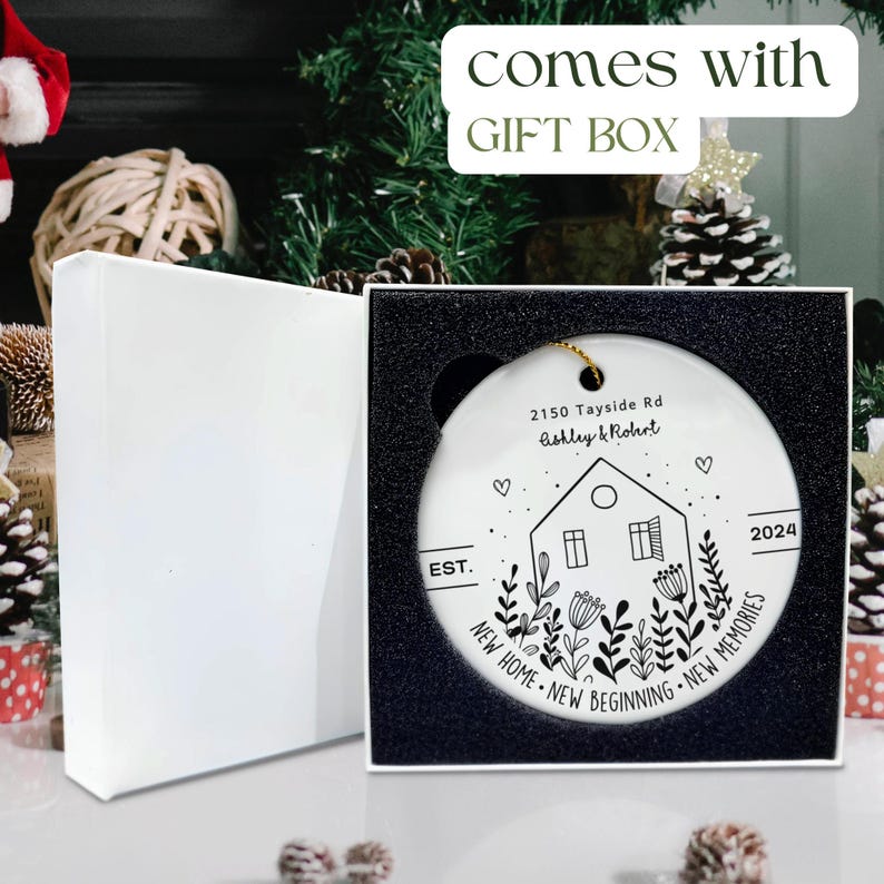 May include: A round white ceramic ornament featuring a black line drawing of a house, flowers, and the text 2150 Tayside Rd, Ashley & Robert, EST. 2024, and NEW HOME NEW BEGINNING NEW MEMORIES, presented in a white gift box with black foam.