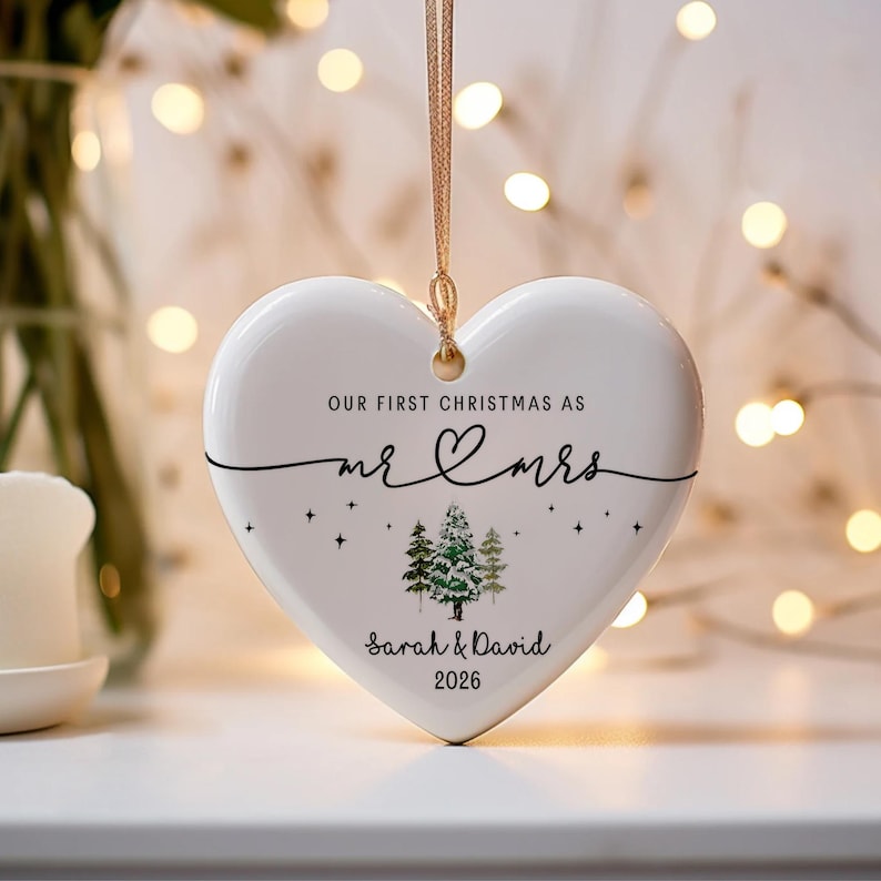 May include: A white heart-shaped Christmas ornament with gold string. The ornament has the text "OUR FIRST CHRISTMAS AS mr & mrs" and the names "Sarah & David" with the year "2026". The ornament also features a small illustration of pine trees.