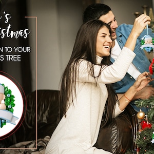 May include: A couple decorates a Christmas tree with blue and green baby ornament. The ornament features a baby with a blue hat and a blue bow. The text "Baby's First Christmas" is visible in the image.