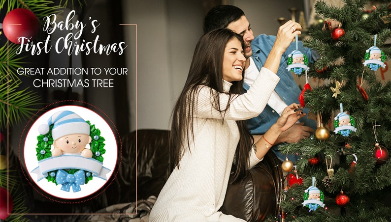 May include: A couple decorates a Christmas tree with blue and green baby ornament. The ornament features a baby with a blue hat and a blue bow. The text "Baby's First Christmas" is visible in the image.