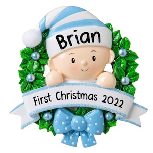 May include: A blue and white personalized Christmas ornament featuring a baby boy peeking over a green holly wreath with a blue bow. The ornament includes the name "Brian" and the text "First Christmas 2022".