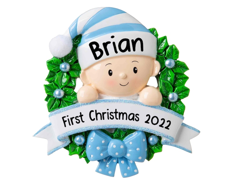 May include: A blue and white personalized Christmas ornament featuring a baby boy peeking over a green holly wreath with a blue bow. The ornament includes the name "Brian" and the text "First Christmas 2022".