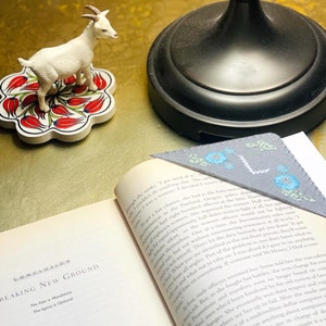 May include: A close-up of an open book with a blue and white embroidered bookmark. The bookmark has a floral design with blue flowers and green leaves. The book is open to a page with the title "Breaking New Ground" and text about discrimination in the workplace.