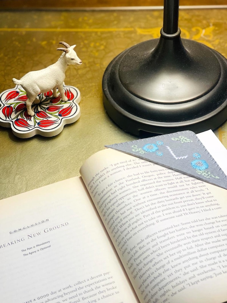May include: A close-up of an open book with a blue and white embroidered bookmark. The bookmark has a floral design with blue flowers and green leaves. The book is open to a page with the title "Breaking New Ground" and text about discrimination in the workplace.
