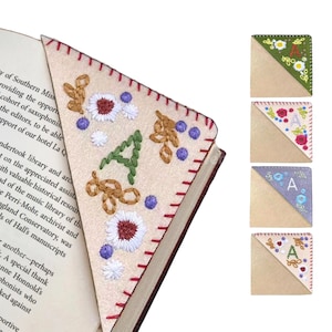 May include: Four handmade felt bookmarks with embroidered flowers and the letter 'A' in different colors. The bookmarks are in the shape of a right triangle with a beige base and a colored top. The colors are green, pink, purple, and beige.