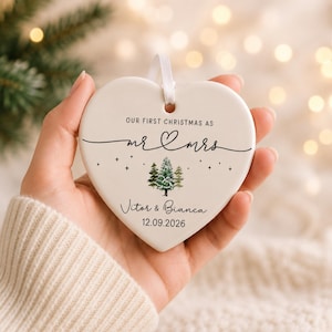 May include: Heart-shaped ceramic Christmas ornament with the text "OUR FIRST CHRISTMAS AS mr & mrs" and a tree illustration. The names "Vitor & Bianca" and the date "12.09.2026" are also printed on the ornament. A white ribbon is attached for hanging.