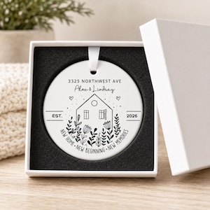 May include: A round white ceramic ornament with a black line art design featuring a house, floral elements, and the text 3325 NORTHWEST AVE, Alex & Lindsay, EST. 2026, and NEW HOME NEW BEGINNING NEW MEMORIES, presented in a white gift box with black foam.