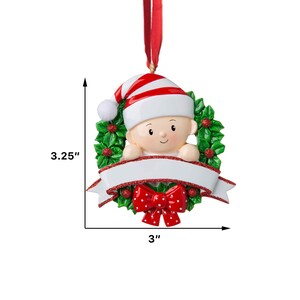 May include: A white and red baby ornament with a red bow and a green holly wreath. The ornament is 3 inches wide and 3.25 inches tall. It has a blank white banner for personalization.