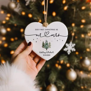 Puede incluir: Adorno navide&ntilde;o blanco en forma de coraz&oacute;n con el texto "Our first Christmas as Mr & Mrs Ashley & Robert 10.10.2024" y una peque&ntilde;a ilustraci&oacute;n de dos &aacute;rboles de hoja perenne.