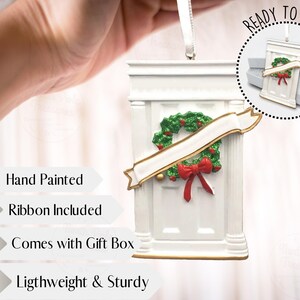 May include: White hand-painted Christmas ornament shaped like a front door with a green wreath and a red bow. The ornament comes with a ribbon and a gift box. "Ready to Gift" is written on the image.
