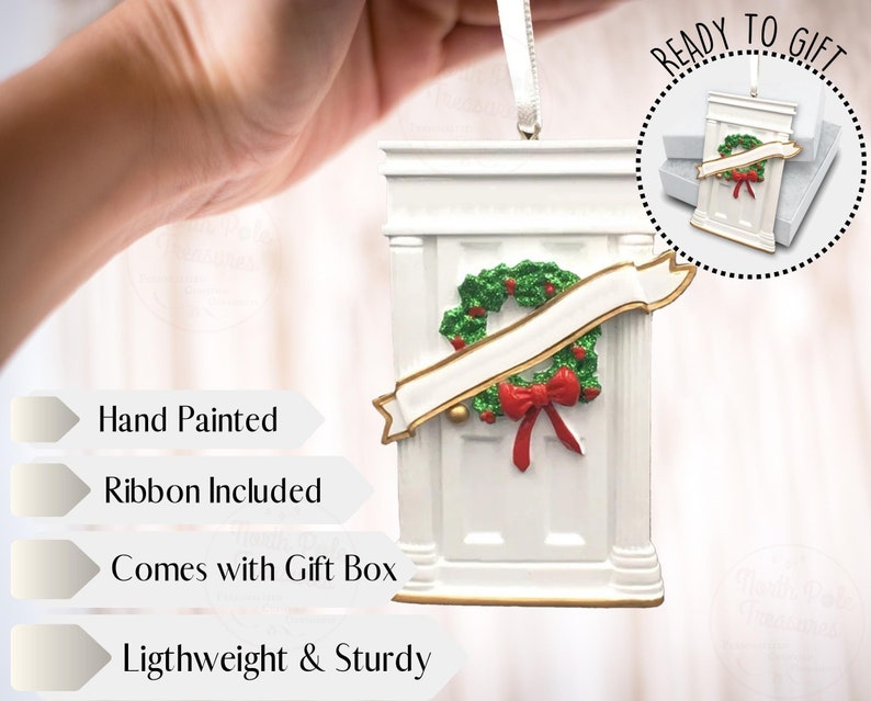 May include: White hand-painted Christmas ornament shaped like a front door with a green wreath and a red bow. The ornament comes with a ribbon and a gift box. "Ready to Gift" is written on the image.