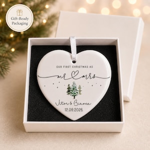 May include: A heart-shaped white ceramic Christmas ornament with the text "OUR FIRST CHRISTMAS AS mr & mrs" and the names Vitor & Bianca, with the date 12.09.2026. The ornament is in a white gift box with a black interior.
