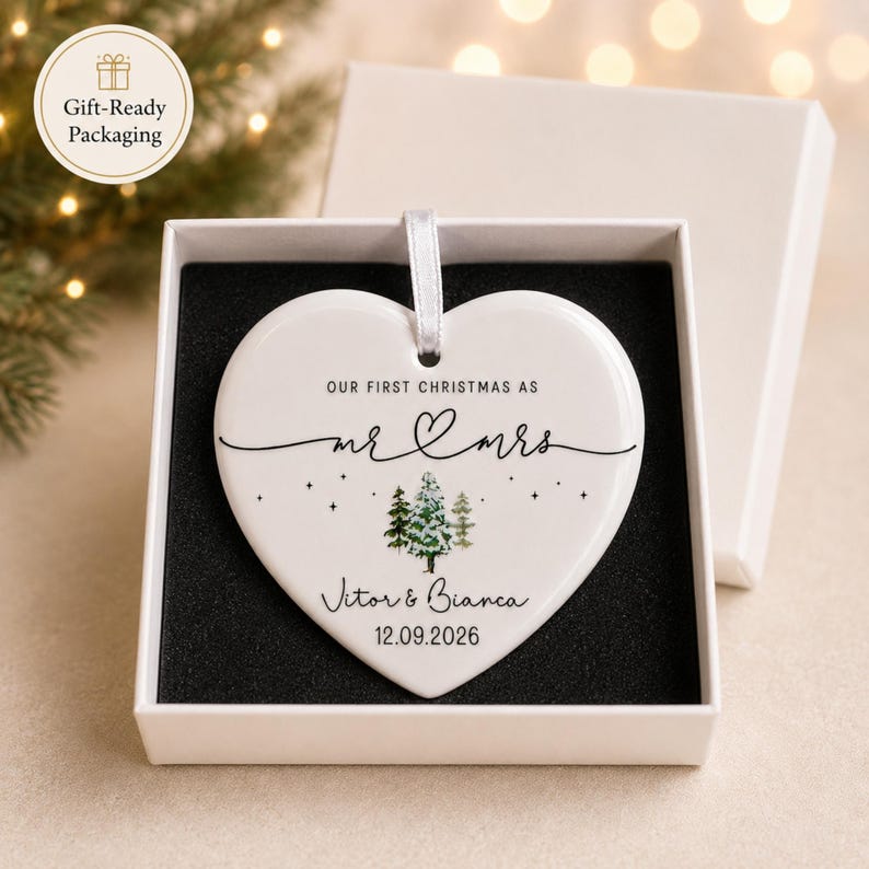 May include: A heart-shaped white ceramic Christmas ornament with the text "OUR FIRST CHRISTMAS AS mr & mrs" and the names Vitor & Bianca, with the date 12.09.2026. The ornament is in a white gift box with a black interior.