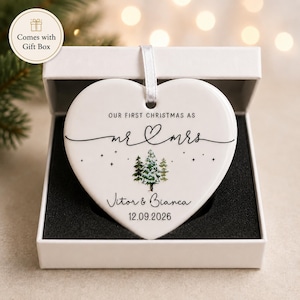 May include: A heart-shaped, white ceramic Christmas ornament with the text "OUR FIRST CHRISTMAS AS mr & mrs" and a forest graphic. The ornament includes the names "Vitor & Bianca" and the date "12.09.2026". It is presented in a white gift box.