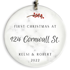 May include: White round Christmas ornament with a marble pattern. The ornament has red berries and a branch on the top. The text on the ornament reads "First Christmas at 124 Cornwall St. KelsI & Robert 2022".