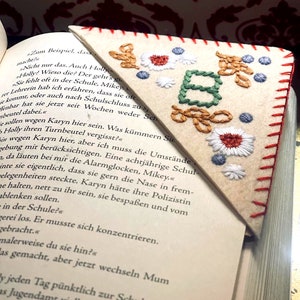May include: A close-up of a book with a handmade bookmark. The bookmark is a triangle shape with a red stitched border. The bookmark is embroidered with the letter "B" in green and brown thread. The background of the bookmark is white felt. The book is open to a page with text in German.