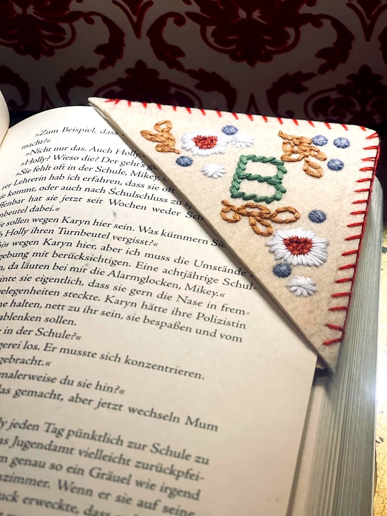 May include: A close-up of a book with a handmade bookmark. The bookmark is a triangle shape with a red stitched border. The bookmark is embroidered with the letter "B" in green and brown thread. The background of the bookmark is white felt. The book is open to a page with text in German.