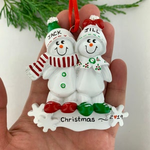 May include: White snowman couple Christmas ornament with red and green accents. The snowman on the left has a red and white striped scarf and the snowman on the right has a red bow. The ornament says "Jack" and "Jill" on the hats and "Christmas 2019" at the bottom.