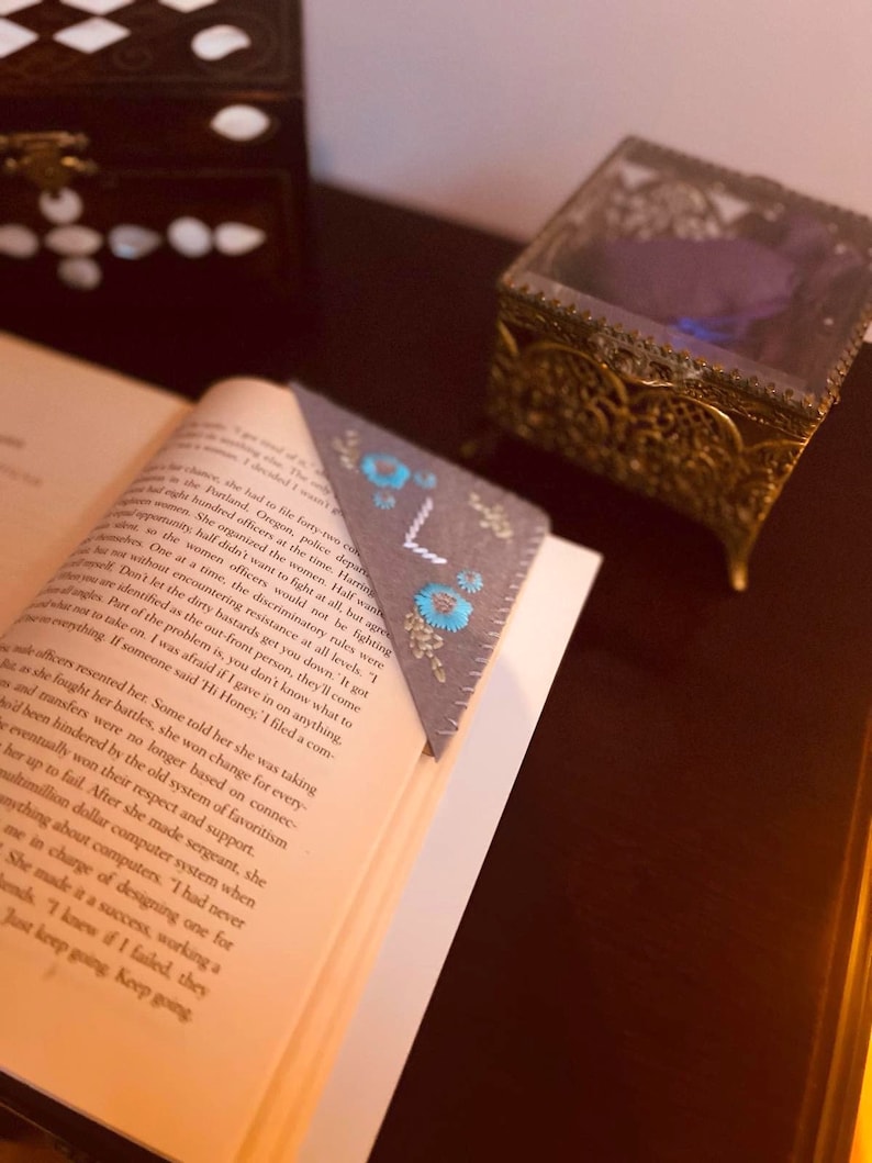 May include: A gray fabric bookmark with blue embroidered flowers. The bookmark is folded and placed on a book with a white cover.
