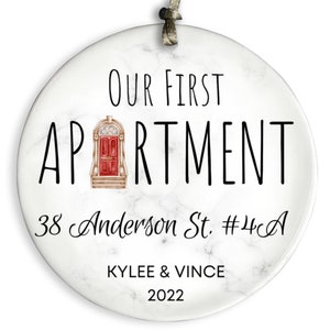 May include: White marble round ornament with black text that reads "Our First Apartment 38 Anderson St. #4C A Kylee & Vince 2022". A small illustration of a red door with a gold frame and steps is in the center of the text.
