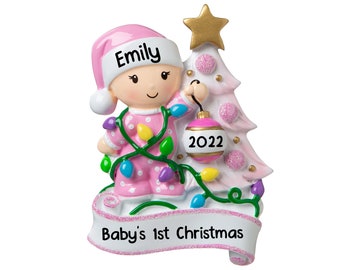 May include: Pink Christmas ornament featuring a baby girl in a pink and white pajamas holding a pink ornament with the year 2022 on it. The ornament also has a pink banner with the text "Baby's 1st Christmas" and the baby's name "Emily" on her hat.