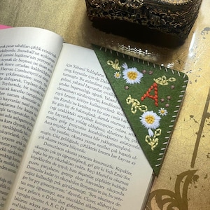 May include: A green felt bookmark with white embroidered daisies and the letter 'A' in orange thread. The bookmark is folded into a triangle shape.