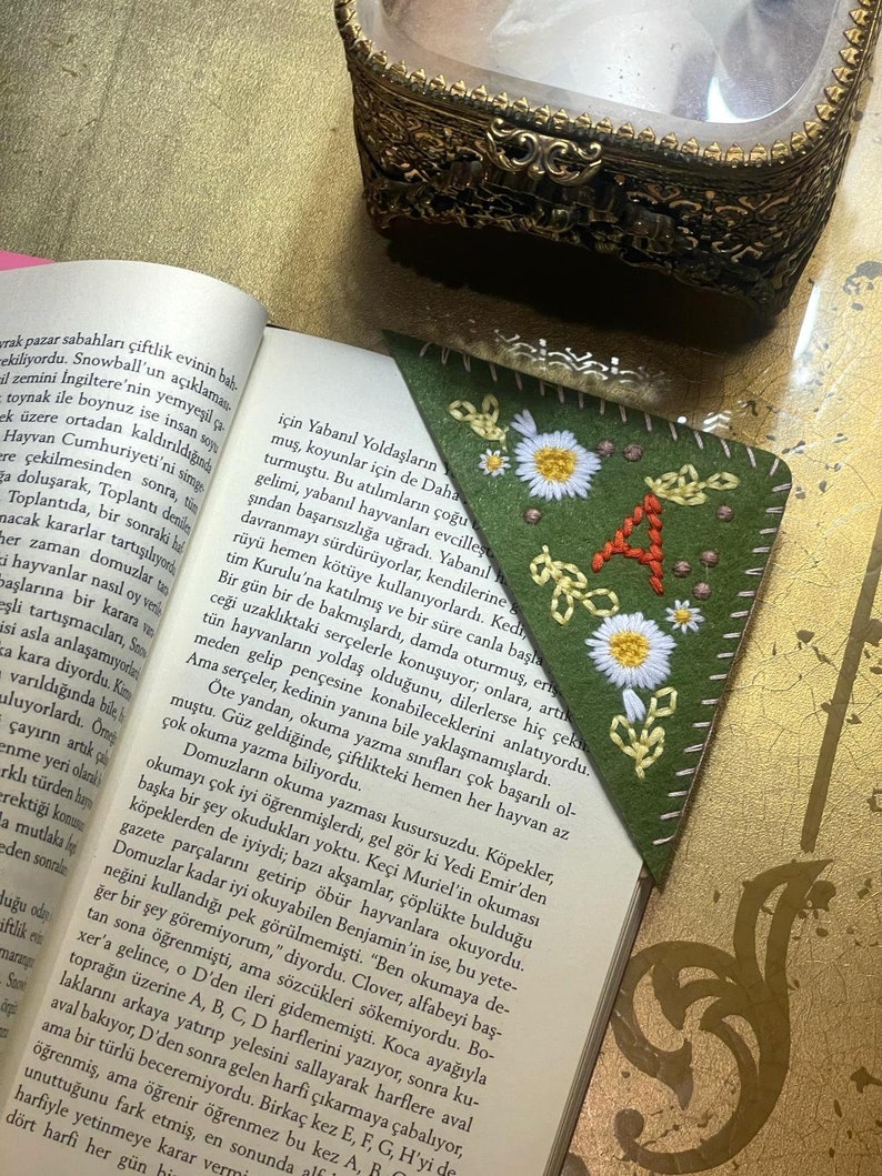 May include: A green felt bookmark with white embroidered daisies and the letter 'A' in orange thread. The bookmark is folded into a triangle shape.