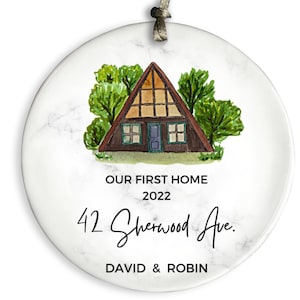 May include: A white round ornament with a marble pattern featuring a watercolor illustration of a brown A-frame house with green trees. The text "Our First Home 2022 42 Sherwood Ave. DAVID & ROBIN" is printed below the house.