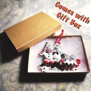 May include: A white snowman family ornament with red and green accents. The ornament features a snowman couple holding hands and a smaller snowman child in a sled. The ornament is in a brown gift box with white filling. The text "Phillips 2019" is on the ornament.