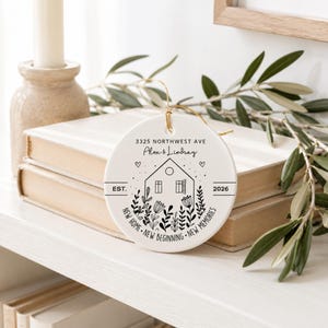 May include: A round white ceramic ornament featuring black line art of a house surrounded by flowers. The text reads 3325 NORTHWEST AVE, Alex & Lindsay, EST. 2026, and NEW HOME NEW BEGINNING NEW MEMORIES. It hangs from a thin gold string.