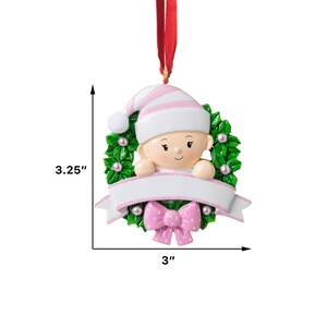 May include: A pink and white baby ornament with a green holly wreath and a blank banner for personalization. The ornament is 3 inches wide and 3.25 inches tall.