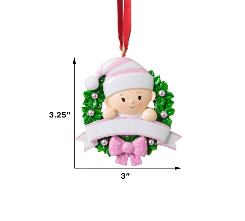 May include: A pink and white baby ornament with a green holly wreath and a blank banner for personalization. The ornament is 3 inches wide and 3.25 inches tall.