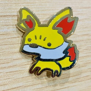 May include: A yellow, red, white, and gold enamel pin featuring a cartoon fox design. The pin has a gold outline and is set against a light wood grain surface. The fox has large ears and a fluffy tail.