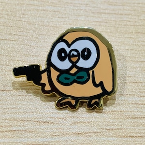 May include: A gold-toned metal enamel pin featuring a tan, round owl character with large white eyes, a small beak, and a green bowtie. The owl is depicted holding a black handgun in its wing, outlined in black with a polished metallic finish.
