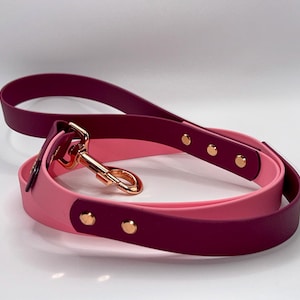 May include: A two-toned dog lead featuring a deep burgundy and light pink strap made of a smooth, flexible material. The lead includes a shiny rose gold metal swivel snap hook and matching rose gold rivets securing the strap segments together.