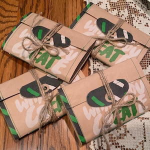 May include: Four beige paper packages with green and black trim, tied with twine. Each package has the words "WITH EAT" in white and green. A dark gray and green oval-shaped closure is on each package.